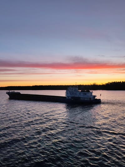 A barge navigates a body of water near Vasil'evskoe, Russia under a vibrant sunset. The sky exhibits bands of color, ranging from purple and blue at the top to pink and orange closer to the horizon. Silhouetted trees line the distant shore. The water displays ripples and reflections of the sky's colors.
