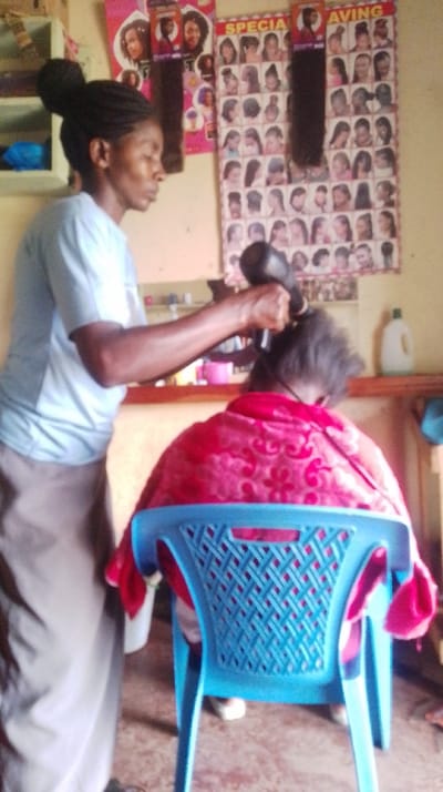 An interior shot in Siaya Township ward, Kenya, depicts a hair stylist actively blow-drying a client's hair. The stylist, a female with braided hair styled into a bun, stands on the left, wearing a light-colored t-shirt and a light brown skirt. She holds a black hairdryer in her right hand, directing it towards the back of the client's head. The client is seated, facing away from the camera, draped in a red cape with a darker patterned design. The client occupies a light blue plastic chair featuring a woven backrest.
The background consists of a light-colored wall adorned with several posters advertising various hairstyles and hair extensions, some with visible text fragments like "SPECIAL" and "AVING." Dark hair extension samples hang from clips on these posters. To the left, a small, light-colored wall shelf holds a blue plastic item. Behind the client, a shelf or counter displays a pink cup and a white plastic bottle with a green cap. The floor is a speckled, light-colored surface.