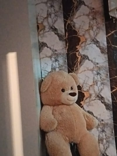 A tan, plush teddy bear sits propped against a wall with a marbled pattern. The bear is facing left, with its head slightly turned towards the viewer. It has black button eyes and a black stitched nose and mouth, giving it a smiling expression. The bear's body is large and rounded, with its arms resting on its lap. A vertical beam of light falls across the left side of the image, illuminating a portion of the wall and the bear's side. The background consists of vertical stripes of a black and white marbled pattern, with accents of gold. The scene appears to be indoors, possibly in a home in Sadisko Pole, Bulgaria.