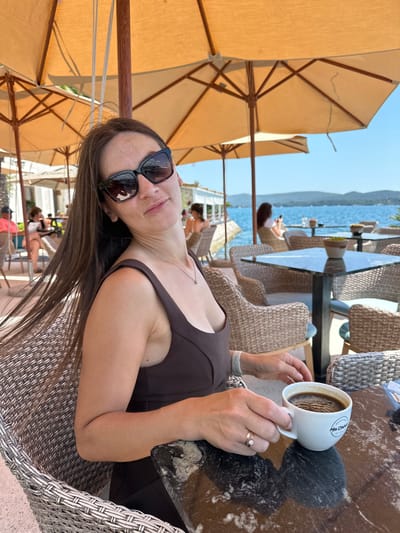 A woman sits at a table outside a cafe, holding a white mug of coffee. She is wearing a dark brown dress and black sunglasses. The cafe is located by the sea in Donja Lastva, Montenegro, with umbrellas providing shade. In the background, other patrons are visible, along with tables and chairs. The water is blue, and mountains are visible in the distance.