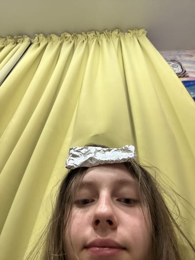 A close-up, low-angle, selfie-style shot shows a young person with light brown hair and fair skin, facing the camera. A crumpled piece of aluminum foil rests on top of their head. The person's eyebrows are furrowed, and their eyes are looking slightly to the left. Their lips are slightly parted. The background consists of yellow curtains, heavily creased and gathered at the top, with a suggestion of a painting or mural visible on the far right. The image appears to be taken indoors. There is no identifiable information suggesting a location in the city of ЖК «Борисоглебское», Russia.