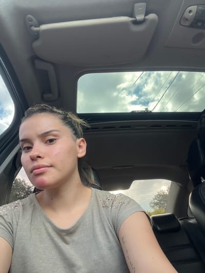 A young woman is photographed inside a car with a sunroof. The sky with clouds is visible through the sunroof. The woman has her hair pulled back and is wearing a gray shirt. She has a tattoo on her upper arm. The interior of the car is primarily gray and black. The image provides no information to verify its location in San Isidro, Argentina.