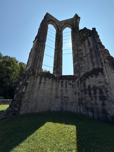 The image shows the ruins of the Walkenried Abbey in Walkenried, Germany. The ruins are made of large stone blocks and appear to be ancient. Two large arched windows are visible, with thin metal bars stretching across them. The sky is clear and blue, and the sun is shining brightly through the windows, creating a lens flare effect. The ruins are surrounded by green grass, and trees can be seen in the background. A fence is also visible to the left of the ruins. The image is taken from a low angle, looking up at the ruins. The overall impression is one of grandeur and decay.