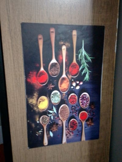 The image is a close-up, slightly angled shot of a framed decorative print featuring an assortment of spices in wooden spoons and small bowls. The print is set against a dark, textured background, likely a wooden surface, with some bokeh effects creating a blurred atmosphere around the spices. 

The arrangement includes several wooden spoons, each holding a different spice. From top left, going clockwise: a spoon with red powder (likely paprika or chili powder), followed by a spoon with brown powder (perhaps cinnamon or ground coffee), then a spoon with dark brown specks (possibly ground pepper or a spice blend). Below these, a spoon holds a light brown powder, and another contains a dark, granular spice. There are also small round bowls filled with yellow powder (turmeric), green herbs (like dried parsley or dill), red peppercorns, and purple peppercorns. A sprig of rosemary is visible towards the top right. Scattered amongst the spices are whole peppercorns and star anise.

The frame of the print is a light brown wood grain. The overall impression is one of culinary richness and visual appeal, common in home decor for kitchens or dining areas.

While the image depicts a decorative print, there is no information within the image itself to suggest its location in Cuiabá, Brazil. The description is therefore limited to the visual content of the print and its immediate surroundings as depicted.