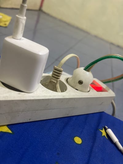 A white power strip is plugged into a wall socket, with a white phone charger and two other plugs inserted into its outlets. One of these plugs has a red indicator light, suggesting it is receiving power. The power strip and plugs are situated on a blue surface adorned with yellow stars and circles, possibly a fabric. In the background, a tiled floor and a wall are visible. The scene appears to be indoors, likely in a residential setting within Anggaswangi, Indonesia.
