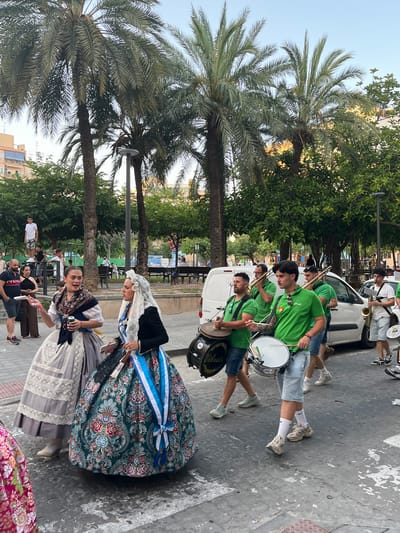 The image depicts a lively outdoor scene in Alicante, Spain, possibly during a festival or parade. Several people are participating in a marching band, dressed in matching bright green t-shirts and casual shorts or trousers. They are playing various instruments, including drums and brass instruments like a saxophone and a trombone. One person is playing a large bass drum.

In the foreground, two women are dressed in traditional Valencian attire. They are wearing elaborate, long skirts with intricate floral patterns and lace details, along with white head coverings and sashes. One woman is holding a fan. The women appear to be smiling or engaged in conversation, suggesting a festive atmosphere.

The background shows palm trees and lush green foliage, characteristic of the Mediterranean climate. There are also buildings and what appears to be a white van, suggesting an urban or semi-urban setting. The lighting suggests it is daytime, likely late afternoon or early evening due to the warm tones. The overall mood is one of celebration and community engagement, with the blend of modern band attire and traditional dress highlighting cultural elements. Some text is visible on the drums, but it is largely illegible due to the resolution and angle.