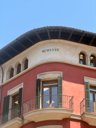 The image shows the upper facade of a building with a red-painted wall and a beige cornice. The cornice is decorated with arched windows and the Roman numerals MCMXXXII, which likely indicate the year 1932. There are balconies with wrought-iron railings. The building is viewed from below, with a clear blue sky in the background. The architectural style appears to be Mediterranean. The building is located in Palma de Mallorca, Spain.