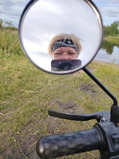 This media file is a selfie taken by a person from their perspective, looking into a motorcycle's rearview mirror. The subject is a person with blonde hair, wearing glasses and a black bandana with a white pattern. They are holding a smartphone, also visible in the mirror, which is capturing the image.
The setting appears to be outdoors, with a body of water and trees reflected in the mirror behind the person. The foreground shows the handlebars of a motorcycle, with the rider's hand on the brake lever. The ground is covered in dry grass and dirt.
The time of day appears to be late afternoon or early evening, given the soft lighting and the sky's appearance, which is partly cloudy. The location is identified as Заямнаўскі сельскі Савет, Belarus, suggesting a rural or semi-rural environment. No text is visible in the image. The overall mood is casual and self-documenting.