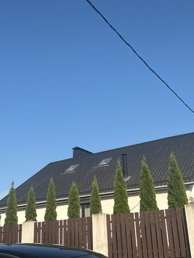 The image shows a close-up view of the side of a modern house with a dark, textured roof and several windows. A brown wooden fence runs along the side of the house, and several tall, cone-shaped evergreen trees are planted in front of the fence. A dark-colored vehicle is partially visible on the right side of the frame. Above the house and fence, a clear blue sky occupies most of the frame, with a thin black wire stretching diagonally across the upper left portion. The lighting suggests it is daytime, likely a sunny day given the clear sky. The scene appears to be residential. There is no visible text in the image.