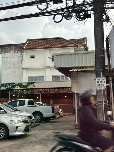 The image captures a street scene in Si Sunthon, Thailand, likely during the daytime given the overcast sky. The foreground is dominated by a tangle of black electrical wires, a common sight in many Asian countries, suspended above a street. To the right of the wires, a multi-story building is visible. The building has a mix of architectural styles, with a reddish-brown tiled roof on the upper section and white facades on the lower levels. Some windows are visible, including small, square ones.
Below the building, a sign for "DADDY'S GRILL" is prominently displayed, suggesting a commercial area with food establishments. The sign features images of food, likely from the grill. Adjacent to the building, several vehicles are parked, including a silver car in the very front and a white pickup truck behind it. The presence of parked cars indicates this is a public area, possibly a road or parking lot adjacent to businesses.
In the background, part of another building or structure is visible, with some signage that is partially obscured and difficult to read clearly, though some text like "TLG-TCH" and "SD-TH-3" can be discerned. There are also blurred figures of people, likely pedestrians or individuals on motorcycles, adding to the dynamic nature of the scene. The overall impression is that of a typical urban street in Thailand, with a blend of residential and commercial elements, and the characteristic overhead network of electrical cables. The weather appears to be cloudy, suggesting it might be a humid day.