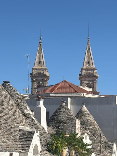This image, taken in the historic Rione Monti district of Alberobello, Italy, captures a scene dominated by the distinctive conical roofs of trulli houses. In the foreground, several trulli with their grey, stacked stone roofs are visible. A white, chimney-like structure rises from one of these trulli. Behind the trulli, a building with a red tiled roof and white walls is partially visible. Towering above everything are two weathered, stone church spires, likely part of the Chiesa di Santa Lucia or the Basilica dei Santi Medici Cosma e Damiano, both iconic landmarks in Alberobello. A slender metal antenna with multiple horizontal elements extends from behind the left spire. The sky is a clear, vibrant blue. No people are visible in the image.
