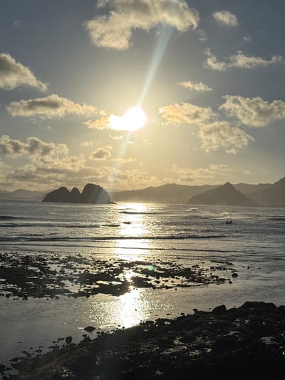 This is a vertical, full shot of Selong Belanak Beach in Selongbelanak, Indonesia, during sunset. The sun is low in the sky, casting a bright glare that dominates the upper half of the image and reflects off the water, creating a shimmering path. Several clouds are scattered across the sky, some illuminated by the sun. In the middle ground, the ocean displays gentle waves, and a small, dark boat is visible on the water. The background features the silhouettes of rolling hills or mountains. The foreground consists of a rocky shoreline, with dark, wet rocks and tide pools reflecting the sunset's light. There are no people visible in the frame.