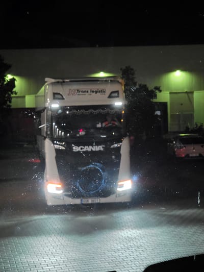 In Zeppelinheim, Germany, at night, a white Scania truck with "JS Trans logistic" written on its side is parked on a paved surface. The truck has its headlights on, illuminating the foreground and the truck itself. The background shows a building with green lights, suggesting it might be a warehouse or industrial facility. There is also a white car with a red tail light parked to the right of the truck. The image appears to be taken from inside another vehicle, as indicated by the windshield and reflections.