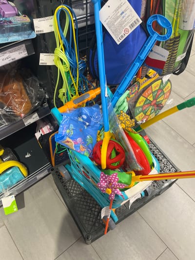 The image shows a collection of children's toys in a shopping cart, possibly in a store. Among the toys visible are jump ropes, a dartboard, a blue bubble wand, and a toy net. The toys are colorful and appear to be new. The shopping cart itself is made of dark gray plastic and has a criss-cross pattern. In the background, shelves are visible with other products, some of which are packaged. A price tag with Russian text is visible on one of the shelves. The floor is tiled with light gray tiles. There are no people visible in the image. The overall scene suggests a retail environment where children's toys are sold. There is no information in the image to suggest it is located in the city of Navahrudak, Belarus.