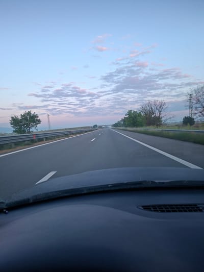 A view from inside a vehicle traveling on a multi-lane highway near Shishmantsi, Bulgaria. The highway has a white dividing line and guardrails on the left side. Trees and vegetation line the side of the highway. The sky is blue with clouds tinged with pink. The interior of the vehicle is visible on the right side of the image.