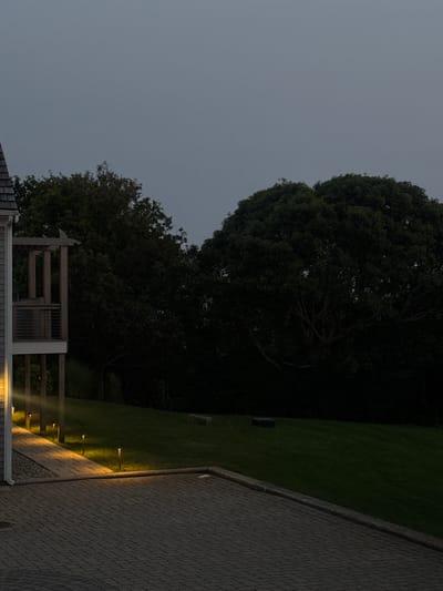 The image depicts an outdoor scene at dusk or early night. A paved walkway, illuminated by a series of small, upright lights, extends from the foreground into the middle ground. The walkway is bordered by a grassy area to the right and a darker, possibly wooded area further back. To the left, a portion of a building, possibly a house or deck, is visible. This building has a grey exterior and a wooden structure, perhaps a porch or balcony, with railings. The sky is overcast and a pale grey. The location is specified as Provincetown, United States of America.