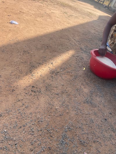 A low-angle, eye-level shot captures a patch of reddish-brown dirt and gravel ground, illuminated by a diagonal beam of sunlight. In the upper left corner of the frame, a crumpled piece of white plastic is visible. Further to the right, a person's arm and hand are partially visible, reaching into a bright red plastic basin filled with white rice. The person is wearing a patterned fabric around their waist. The background is out of focus, showing more of the dirt ground and some faint structures or vegetation in the distance. The scene appears to be outdoors, possibly in a rural or informal urban setting within Wauro Jabbe, Nigeria.