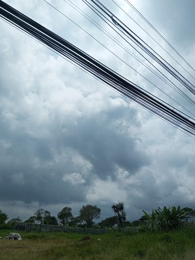 The image captures an outdoor scene in Batulawang, Indonesia, under a heavily overcast sky dominated by dark grey cumulus clouds. A prominent array of black utility cables runs diagonally across the upper portion of the frame, from the upper left to the middle right. The lower foreground consists of an uncultivated area with mixed green and dry grasses. In the midground, a low concrete wall, marked with dark patterns consistent with graffiti, stretches horizontally across the scene. To the immediate left of the foreground, several light-colored, irregular objects, possibly debris or rocks, are present. Behind the wall, a line of trees with varied foliage is visible, including denser leafy trees and a cluster of banana plants on the right. No individuals or discernible actions are observed within the frame.