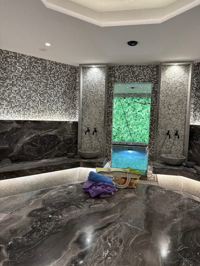 This media file depicts a luxurious indoor swimming pool area, possibly a spa or private residence, in Hisarya, Bulgaria. The scene features a small pool with clear blue water, illuminated from below, reflecting light onto the surrounding mosaic tile walls. One wall displays a large, vibrantly green malachite-patterned panel embedded with small lights, creating a starry effect.
Adjacent to the pool are two shower stalls, each with dark fixtures and stone basins. The area is finished with dark, veined marble countertops and walls, complemented by subtle, integrated lighting that casts a warm glow. On one of the marble surfaces, a straw tote bag rests, containing what appears to be a towel and possibly some personal items. Two folded towels, one blue and one purple, are placed atop the bag.
The overall ambiance is one of tranquility and opulence. The lighting suggests it is either evening or a dimly lit indoor space, enhancing the serene atmosphere. There are no people clearly visible, though the presence of towels and the inviting pool suggests recent or imminent use. The tiling patterns, varying from small mosaics to larger, polished marble, add texture and visual interest to the sophisticated design. The environment appears to be clean and well-maintained, exuding a sense of calm and relaxation.