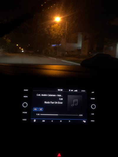 The image shows a car's infotainment system playing "Nada Fue Un Error" by Coti and Andrés Calamaro. The time is 18:58, and the temperature is 23.0°C. Options displayed on the screen include radio, media, phone, voice, app, sound, car, and menu. The song is 1 minute and 36 seconds in, with 1 minute and 51 seconds remaining. Controls for playback are visible. A red hazard light indicator is illuminated. The scene outside the car window is dark with a street light.