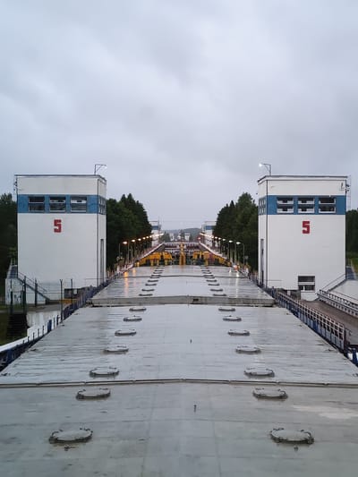 The image captures a view of a canal lock system in Alekseevskoe, Russia. The perspective is from the top of a vessel or structure, looking forward along the canal. On either side, imposing white buildings with blue trim rise vertically. Each building features multiple windows on its upper section and a large red numeral "5" prominently displayed on its facade. A light fixture is visible on top of the building to the right.
The canal itself is flanked by rows of streetlights, which are illuminated, suggesting it may be dusk or a cloudy day. Beyond the immediate canal structures, a dense line of green trees forms a backdrop. In the distance, more of the canal system, including what appear to be yellow and grey machinery and other lock structures, can be seen receding into the haze. The surface in the foreground, presumably the deck of the vessel, is a large, grey, flat expanse marked with numerous circular, lidded access points or hatches arranged in rows. The surface appears wet, reflecting the overcast sky and artificial lights. No people are visible in the image.