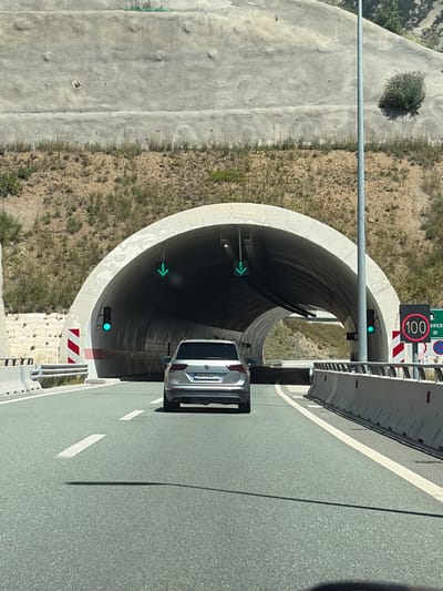 The image captures a view from a vehicle approaching the entrance of a tunnel on a sunny day. The road ahead is asphalt with white lane markings. A silver SUV is in front of the vehicle, driving towards the tunnel. To the left of the road, there are concrete barriers and a red and white striped marker. The tunnel entrance is a large, arched opening in a concrete structure. Green traffic lights are visible, indicating that the lane is open. A speed limit sign displaying "100" in a red circle is to the right of the road, followed by a sign for "Service m" and a "No stopping" sign. The tunnel walls are illuminated. The landscape surrounding the road is a dry, grassy hillside with some rocky areas and concrete retaining walls. The scene is likely a highway leading to or from a tunnel in the Lopate area of Montenegro, given the "Service m" signage which suggests it's a highway service area sign.