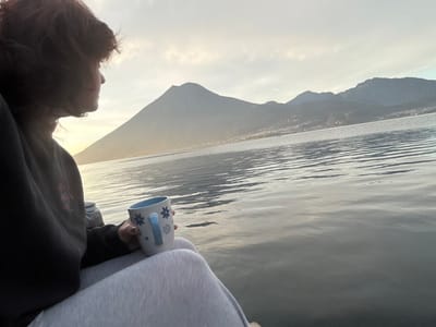 A person with dark, medium-length hair is seated facing right, their upper body partially obscured, wearing a dark long-sleeved top and light gray sweatpants. The individual holds a white ceramic mug, decorated with blue floral patterns and a light blue handle, in their right hand. The scene depicts a view over the calm waters of Lake Atitlán from the city of San Pablo La Laguna, Guatemala. In the background, a prominent conical mountain or volcano, consistent with Volcán San Pedro, rises on the left, with a range of mountains extending to the right. Distant settlements are visible along the far shoreline. The sky presents a soft, pale illumination, suggesting either dawn or dusk, with the light reflecting subtly on the water's surface.