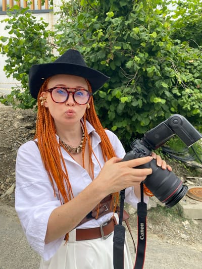 A woman with bright orange braided hair, wearing a black cowboy hat, large tortoiseshell glasses, a white button-up shirt, and white pants, poses for a photo outdoors. She is holding a professional Canon camera with a flash attachment, and has a brown leather belt and a distinctive chunky gold necklace. The background consists of lush green foliage and a gravelly area.