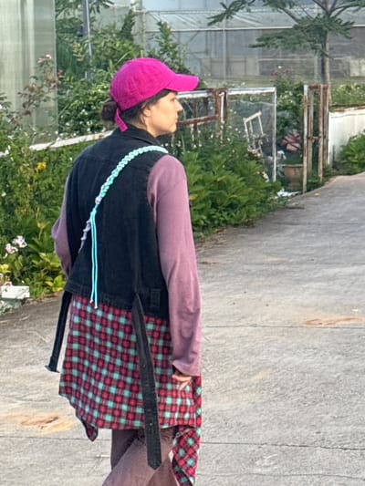 A woman with dark hair pulled back and wearing a pink baseball cap walks away from the camera on a paved path in a garden setting. She is wearing a purple long-sleeved shirt under a dark denim vest, and a red and teal plaid skirt. A light blue and white braided strap is visible across her back, attached to a bag. The path is bordered by lush green foliage and flowering plants, with a greenhouse structure visible in the background. The scene is consistent with the agricultural and floral environment found in Lam Vien - Da Lat, Vietnam.