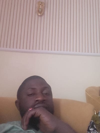 The image depicts an indoor scene in Babawa, Nigeria. A dark-skinned adult male is positioned in the foreground, shown from the chest up. He has short dark hair and a beard, wearing a light green short-sleeved shirt. His left hand is raised to his lower face, with fingers resting near his chin and mouth; a red-stoned ring is visible on his left ring finger. He looks directly towards the camera. To his right, a mustard-yellow upholstered sofa arm is partially visible. The background consists of a plain, light-colored wall in the mid-ground, surmounted by a white wall section featuring vertical ridged paneling or wallpaper. A small, brass-colored wall fixture, possibly a light or decorative element, is mounted on this ridged section in the upper center.