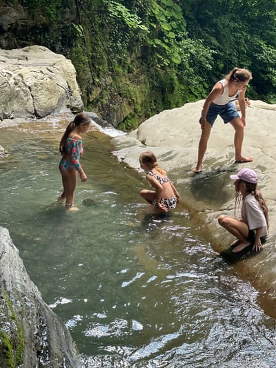 The image depicts three young girls in a natural setting with water and rocks. One girl, in the foreground, is wearing a colorful swimsuit and standing in shallow, clear water. She has long, dark hair and her left arm is bent at the elbow. Behind her, another girl in a leopard-print bikini is crouching in the water, looking downwards. Her hair is light brown and tied back. In the background, a third girl, wearing a white top, blue shorts, and a pink baseball cap, is standing on a large, light-colored rock. She is looking towards the other two girls. Her hair is light brown and pulled back into a ponytail. The surrounding environment consists of lush green foliage and large, weathered rocks, suggesting a natural, possibly wooded area with a stream or waterfall. The water is clear and reflects the light. The overall impression is one of a sunny day, with the girls enjoying an outdoor activity. There is no visible text in the image.