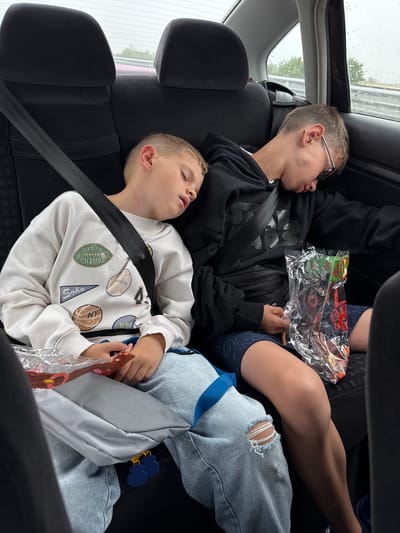 Two young males are asleep in the back seat of a vehicle. The male on the left is wearing a white sweatshirt with various patches and light-wash ripped jeans. He is resting his head on a gray fabric object. The male on the right is wearing a black hoodie and dark patterned shorts. He has glasses on and is holding a clear plastic bag containing what appears to be candy. Both males are wearing seatbelts. The background shows the interior of a car, with the rear seats and headrests visible. Through the window, a blurry, overcast sky and some greenery can be seen, suggesting it is daytime and possibly raining. The scene is consistent with individuals sleeping during a car journey. The image does not contain any specific visual cues that would place it in Malo Konare, Bulgaria.