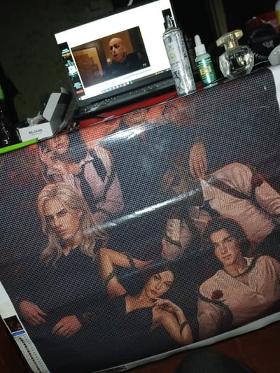 The media file shows a diamond painting kit spread out on a dark surface. The diamond painting itself depicts a group of people, appearing to be a couple or close friends, in what looks like a studio setting with dark backgrounds and hints of warm lighting. The individuals are positioned close together, with one person leaning in towards another, suggesting intimacy or a shared moment. The style of the painting is detailed, with small, colored diamond-shaped beads forming the image.
To the left of the diamond painting, a laptop is open, displaying a picture of a bald man with a serious expression. Scattered around the laptop and the painting are several objects, including a clear glass perfume bottle with a gold cap, a small green bottle with a white cap, and a spray bottle with a decorative pattern. There is also a small white box with green and black text. A colored legend or key for the diamond painting is visible along the bottom edge.
The overall impression is of a craft project in progress, with the artist's tools and the canvas laid out. The lighting in the scene is dim, suggesting it might be nighttime or an indoor setting with artificial light. The emotions conveyed by the painted subjects appear to be calm and perhaps contemplative. The location context of Chutove, Ukraine, is provided, but there are no specific visual cues in the image to confirm this location.
Visible text includes "RE CARE" on the white box, and partial text on the spray bottle and the laptop screen, though it is largely illegible. The diamond painting itself has a printed legend with color codes and symbols.