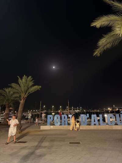 The image depicts an outdoor scene at night in Port d'Alcúdia, Spain. A large sign spelling out "PORT D'ALCÚDIA" in blue, three-dimensional letters is prominently displayed. Several palm trees are visible, with their fronds silhouetted against the dark sky. The moon is visible in the sky, and there are streetlights illuminating the area.
In the foreground, a young person wearing a white outfit and sandals is standing and looking to their left. To the right of the sign, a woman in a yellow patterned outfit and a young child in a yellow dress are walking away from the camera. Other people are present in the background, near the sign and along the waterfront, which appears to be a marina with several boats docked. The ground is paved with patterned tiles.