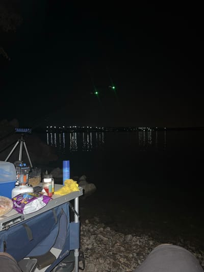 The image depicts a nighttime scene at the shore of a body of water. In the foreground, a portable table is set up with various items. These include a blue cooler, a small portable stove, jars of what appear to be food or bait, a bag of chips, and a yellow cloth. A fishing rod is propped up on a tripod in front of the table. The water reflects lights from a distant shoreline, which appears to be populated with structures or lights. Two green lights are visible in the sky, possibly indicating a distance object or light source. The ground is covered with rocks and pebbles. The scene suggests an outdoor activity, likely fishing, taking place at night. There is no visible indication of the city of Voloske, Ukraine.