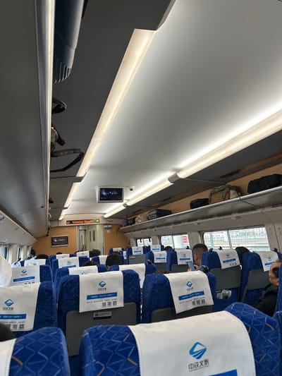The scene depicts the interior of a passenger train carriage, located within Huangdao District, China. The perspective is longitudinal, looking rearward down the aisle from an elevated point within the front section of the carriage. The interior is brightly illuminated by overhead fluorescent strip lights and ambient light entering through windows on both sides. The white ceiling incorporates ventilation grills and integrated overhead luggage compartments. These compartments contain various personal items, including backpacks, a camouflage-patterned bag, and several hard-sided suitcases. Two small, rectangular monitors are mounted to the ceiling, displaying indistinct visual content, and a dome-shaped security camera is also affixed to the ceiling near the monitors. Further down the aisle, an electronic information display panel is visible, partially showing "tination Shangha" and a restroom icon. The blue fabric seats are arranged in multiple rows, each equipped with white headrest covers featuring a blue logo and QR codes. Numerous passengers are seated; most are viewed from behind. In the midground, a male passenger with short, dark hair is observed holding a mobile phone. The exterior visible through the windows suggests an urban environment with elevated infrastructure.