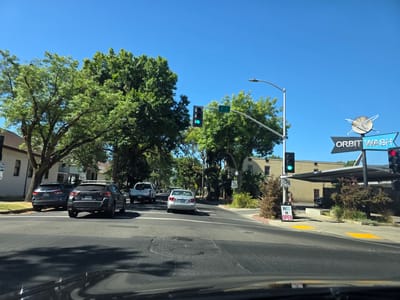 This media file captures a bright, sunny day in Sacramento, California, likely during midday given the strong shadows and clear blue sky. The scene is a street intersection with multiple cars, trees lining the road, and buildings in the background.
In the foreground, the perspective is from inside a vehicle, showing the dashboard and windshield. Several cars are visible on the road. A dark SUV with license plate AXOR076 is in the left lane, while a lighter-colored sedan is ahead in the right lane. Further down the road, a white pickup truck is also visible.
Traffic signals are prominent, with a green light illuminated, indicating traffic can proceed. A street sign indicates "T ST" and another reads "SPEED LIMIT 30". To the right, a sign for "ORBIT WASH" with a circular, atomic-like logo is clearly visible, along with a red traffic signal. A sign on the sidewalk states "IS OPEN".
The presence of mature trees suggests a well-established neighborhood. The overall impression is a typical urban street scene on a clear, warm day. There are no people clearly visible in the image. The focus is on the road, vehicles, and surrounding infrastructure.