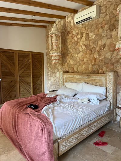 A bedroom in a rustic, possibly Mediterranean-style dwelling features a bed with white linens and a crumpled pink throw blanket. A black item with white lettering, possibly a pair of slippers or a small bag, rests on the pink blanket. A small, white stuffed animal is on the bed. The headboard of the bed is intricately carved wood. The walls are made of large, irregular stones. A modern air conditioning unit is mounted high on one stone wall. A decorative hanging lamp with shell-like ornaments hangs from the ceiling. To the left of the bed are large wooden closet doors with a geometric slatted design. Red flip-flops are on the tiled floor. The image appears to be taken from a slightly elevated angle, looking down into the room.