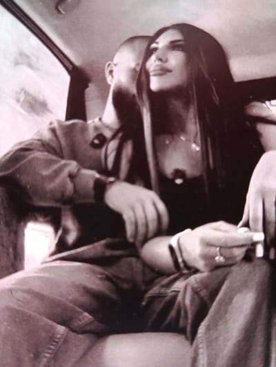 The black and white image shows a close-up view of a man and a woman sitting together in a vehicle, possibly a car. The man, who is bald, is positioned behind and to the left of the woman. He is wearing a long-sleeved shirt and has his left arm resting on his leg. The woman, with long dark hair, is looking to her right, away from the camera. She is wearing a dark top with a necklace. Her hands are visible in the foreground, one holding what appears to be a cigarette. The interior of the vehicle is dimly lit, with some light coming in from a window on the left. The image is grainy, suggesting it was taken with a low-quality camera or in low light conditions. There is no discernible information in the image to indicate a specific location in the city of Bălți, Moldova.