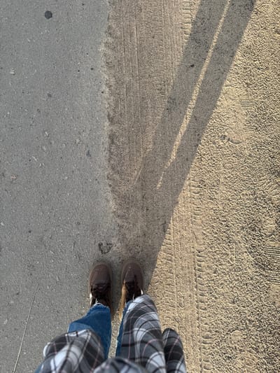 This is a high-angle, first-person perspective shot of a person's lower legs and feet standing on a textured surface that appears to be a dirt or gravel path. The path is divided into two distinct sections: the left side is a darker, smoother asphalt surface, while the right side is a lighter, rougher material with visible tire tracks and imprints of what appear to be footprints. A shadow, cast from an unseen object, stretches diagonally across the lighter portion of the path. The person is wearing blue jeans and brown lace-up shoes. A plaid shirt is visible on the lower legs, suggesting the person is wearing a checkered shirt or pants. A thin, metallic object, possibly a stick or a long nail, lies on the asphalt to the left of the person's feet. The lighting suggests it is daytime, likely late afternoon due to the long shadow. The scene is devoid of any urban context that would specifically place it in Rossosh, Russia; the environment appears to be a common outdoor pathway found in many locations.