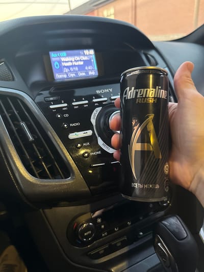 In this rotated image, a person's hand is holding a can of Adrenaline Rush energy drink. The can is black with silver and gold lettering that says "Adrenaline RUSH" and "ВСЕ ТЫ МОЖЕШЬ" (which translates to "You Can Do Anything"). The background appears to be the interior of a car, specifically the dashboard area. A car stereo system is visible with buttons and a screen displaying information. The screen shows the time as 19:45 and lists a song title "Walking On Chươn" with the artist "Heath Wunter". There are also Russian text entries, "Др. 6/18 4:40" and "Повтор Олуч. Окан. Ишф."
The scene suggests someone is in their car, possibly listening to music or reviewing their playlist, while holding an energy drink. The lighting and the interior of the car indicate it is likely evening or night. The image is taken from a close-up perspective, focusing on the hand, the energy drink can, and a portion of the car's dashboard.