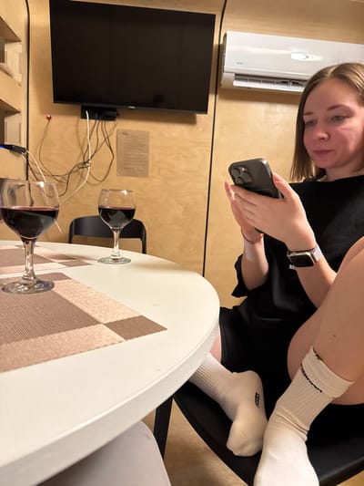 An indoor scene depicts a person seated at a white, round table, using a smartphone. The individual, with light brown hair, wears a black top or dress, white ankle socks (one marked "REESS" on the sole, the other "COMMEN" on the ankle), a grey smartwatch, a multi-colored beaded bracelet, and a small white bandage on the left forearm. Two stemmed wine glasses, each containing red liquid, are positioned on the table; one sits on a beige and brown checkered placemat. In the background, a flat-screen television is mounted on a light-colored wooden wall, with visible cables and a small shelf underneath. To the right of the television, an air conditioning unit is mounted. A piece of paper with text is affixed to the wooden wall. A black chair is partially visible behind the table. The specific location within the city of Vasil'evo, Russia, is not discernible from the provided image details.