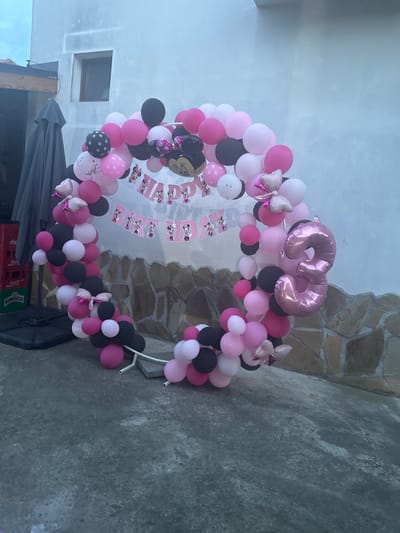 The image shows a balloon arch in a courtyard setting, possibly in the city of Tryavna, Bulgaria. The arch is circular and made of pink, black, and white balloons, with some balloons featuring polka dot patterns. It is decorated with Minnie Mouse cutouts and bows. A large pink foil balloon in the shape of the number "3" is prominently featured on the right side of the arch. In the background, there is a white building with a small window, a stone wall, and some stacked crates. The ground is paved with concrete, and there are some grey umbrella structures on the left side of the image. No people are visible in the scene.