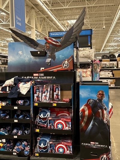 The image captures a brightly lit retail store aisle featuring a prominent promotional display for Marvel's Captain America. The setting is typical of a large department store or supermarket in Worcester, United States, with overhead fluorescent lighting and a white ceiling with exposed metal framework.

The central subjects are two large cardboard displays for "Captain America: Brave New World." On the left, a life-sized standee depicts Sam Wilson as Captain America, complete with his large, spread wings and the iconic vibranium shield, set against a blue sky background. To its right, a tall display box features an illustration of Sam Wilson in his Captain America suit, also holding the shield.

Below these displays, a variety of Captain America-themed merchandise is neatly arranged on shelves and in bins. These items include small boxed action figures or toys, toy Captain America shields, and blue Captain America helmets. Further down, there are Captain America-themed apparel items, such as t-shirts and shorts, alongside blue sandals adorned with the Captain America star-and-shield emblem. Some product tags show prices, such as "$9.98."

In the background, other retail shelves hold various products, including patterned pillows and other packaged goods. A blue hanging sign on the left reads "Home Goods," indicating the section of the store. Another blue sign above the standee partially reads "Everyday Low Price." Key visible text on the displays includes "MARVEL STUDIOS CAPTAIN AMERICA NEW WORLD" on the winged standee and "MARVEL STUDIOS CAPTAIN AMERICA BRAVE NEW WORLD" on the larger display box, promoting the upcoming film. A security camera is visible mounted on the ceiling structure.
