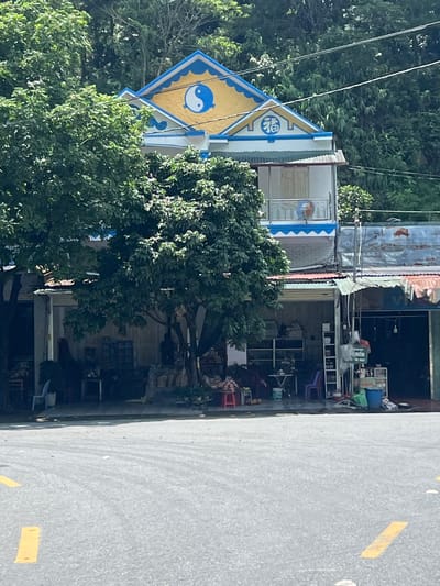 A street-level, eye-level, medium shot captures a multi-story building with a distinctive yellow and blue roof adorned with a Yin and Yang symbol and a blue Chinese character on its facade. The building is situated in Binh Lu, Vietnam, evidenced by a sign in Vietnamese reading "XÃ BÌNH LƯ" above a shop entrance. The ground floor of the building features open-fronted shops, with tables, chairs, and shelves visible within. Lush green trees partially obscure the view of the shops. The foreground shows a paved road with yellow painted lane markers. The overall scene depicts a commercial area, likely a marketplace or a street with small businesses.