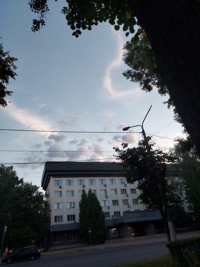 The image shows a building in Zheleznogorsk, Russia, with a street and trees in the foreground. The building is a multi-story structure with a light-colored facade and numerous windows. A streetlamp is visible to the right of the building, and trees line the street and surround the building. A car is parked on the side of the road. The sky is partly cloudy, with a few wispy clouds against a pale blue background. The overall impression is that of a typical urban scene in the late afternoon or early evening.