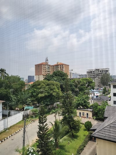 The image displays an elevated view of Falomo, Nigeria, under a partly cloudy sky. In the immediate foreground, the dark-tiled roof and a section of a light-colored building are visible on the right. Adjacent to this is a green lawn featuring manicured shrubs and several trees, including two tall coniferous trees and a palm. A small, single-story utility building with a dark roof and two front windows stands on the lawn. A paved road, bordered by black and white striped curbs, curves through the scene, with utility poles and overhead wires positioned alongside it.

To the left of the road, behind a white perimeter wall, a light blue swimming pool is visible, surrounded by paved areas and tropical foliage, including a large fan-shaped plant. The midground consists of a dense canopy of various green trees. In the background, several multi-story buildings dominate the skyline. Two prominent light-brown buildings with reddish-brown roofs are visible, one featuring a large metal communication tower on its rooftop. To the right, a multi-story concrete structure is under construction, revealing exposed framework and scaffolding. Additional high-rise buildings, some with reflective facades, are discernible on the distant horizon amidst further patches of greenery. No people or vehicles are visible in the scene.
