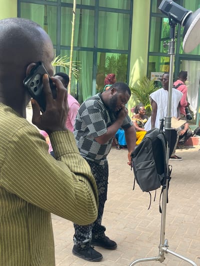 An outdoor scene in Madina, Ghana, depicts multiple individuals gathered on a paved area, likely during a production or event. In the immediate foreground, the back and right side of a dark-skinned man wearing a vertical olive-green striped long-sleeved shirt are visible; he holds a dark smartphone with multiple rear cameras to his right ear, engaged in a call. Adjacent to him, a dark-skinned man with a full beard wears a black and white checkered short-sleeved shirt and dark patterned trousers, possibly with a logo print; he also holds a phone to his left ear, looking downwards.
To the right, a tall, silver-colored light stand with a flash head (labeled "AD600BM") and a softbox reflector is positioned. A dark grey backpack hangs from this stand. Behind the lighting equipment, a dark-skinned man wearing a loose-fitting white sleeveless garment and glasses stands, looking towards the right. He appears to be holding papers or a clipboard.
In the mid-ground and background, additional individuals are present, including a person in a pink shirt, a woman with reddish hair, a woman in a yellow top and blue skirt, and a person holding a camera, suggesting a film or photography set. The background features a modern building with large, green-tinted glass windows, reflecting ambient light. A white sign with red text, partially visible through the glass, reads "NO FACE MA ENZ." The ground is laid with interlocking reddish-brown pavers.
