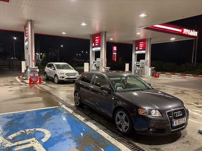 At night, two cars are parked at a Lukoil gas station in Lozen, Bulgaria. A dark gray Audi A3 is in the foreground, parked next to a gas pump. The car's front is visible, showing its license plate "BG A 0215 HC." A white Ford SUV is parked further back, to the left of the Audi, with its license plate "CB 7325 BE" clearly visible.
The gas station's canopy is lit by bright ceiling lights. There are multiple gas pumps visible, with signs indicating fuel types like "ecto 100," "ecto," "A-95H," and "Diesel." The ground is wet, suggesting recent rain or cleaning. A blue parking spot with a faded white wheelchair symbol is in the lower left corner of the image. The overall atmosphere is functional and typical of a nighttime gas station scene. The visible text includes brand names like "Lukoil" and fuel types, along with Bulgarian text that appears to be "Innovative Fuels."
