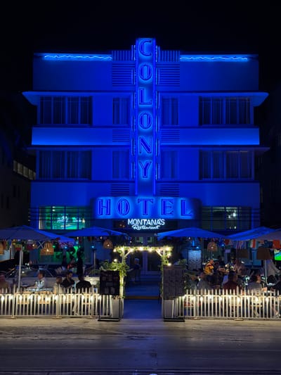 This image depicts the exterior of a hotel and restaurant at night, likely in Miami Beach, United States. The dominant subject is a building illuminated with vibrant blue neon lights, displaying the words "COLONY HOTEL" and "MONTANAS Restaurant" in neon lettering. The hotel's facade is a blend of modern architectural lines with Art Deco influences, characterized by its vertical elements and horizontal window bands.
In front of the hotel, an outdoor dining area is set up, with tables and chairs occupied by several people. The people appear to be patrons of the restaurant, engaged in conversations and dining. The atmosphere seems relaxed and lively, with string lights illuminating the outdoor seating area, creating a warm and inviting ambiance. The time of day is clearly night, as indicated by the darkness of the sky and the artificial lighting.
The foreground shows a paved area, likely a sidewalk or street, adjacent to the restaurant's seating. White fencing encloses the dining space. Various flags are displayed on poles, adding a decorative element to the scene. Visible text includes the names of the hotel and restaurant, as well as what appear to be menus displayed on stands. The overall scene suggests a typical evening at a popular beachfront establishment, with a mix of architectural style, dining activity, and nocturnal illumination.